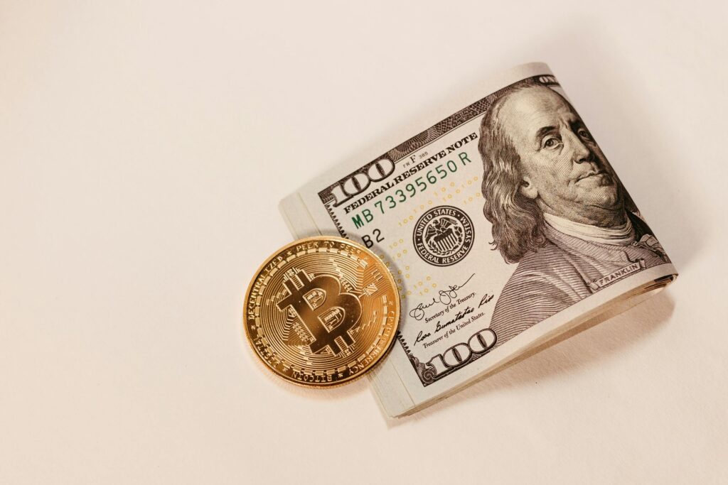 A Bitcoin coin placed next to a rolled US hundred dollar bill, symbolizing cryptocurrency and traditional currency.