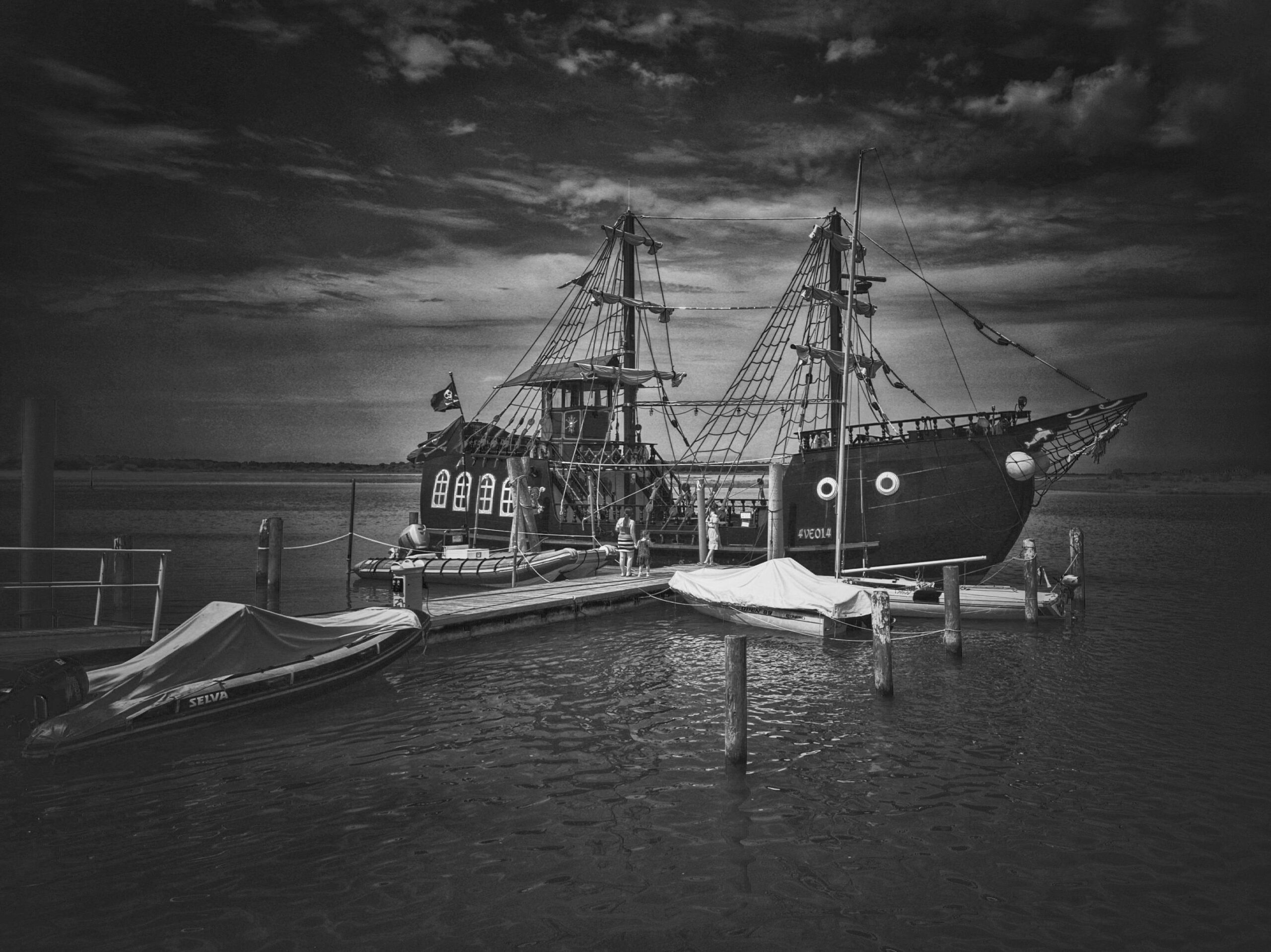 Grayscale image of a pirate ship docked in a peaceful harbor setting.